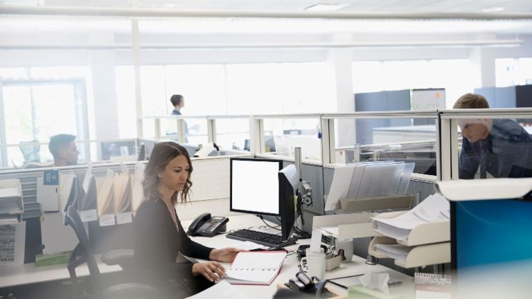 Дигитализацията: трима служители са в отворен офис – една жена е седнала на компютъра си измежду хартиите