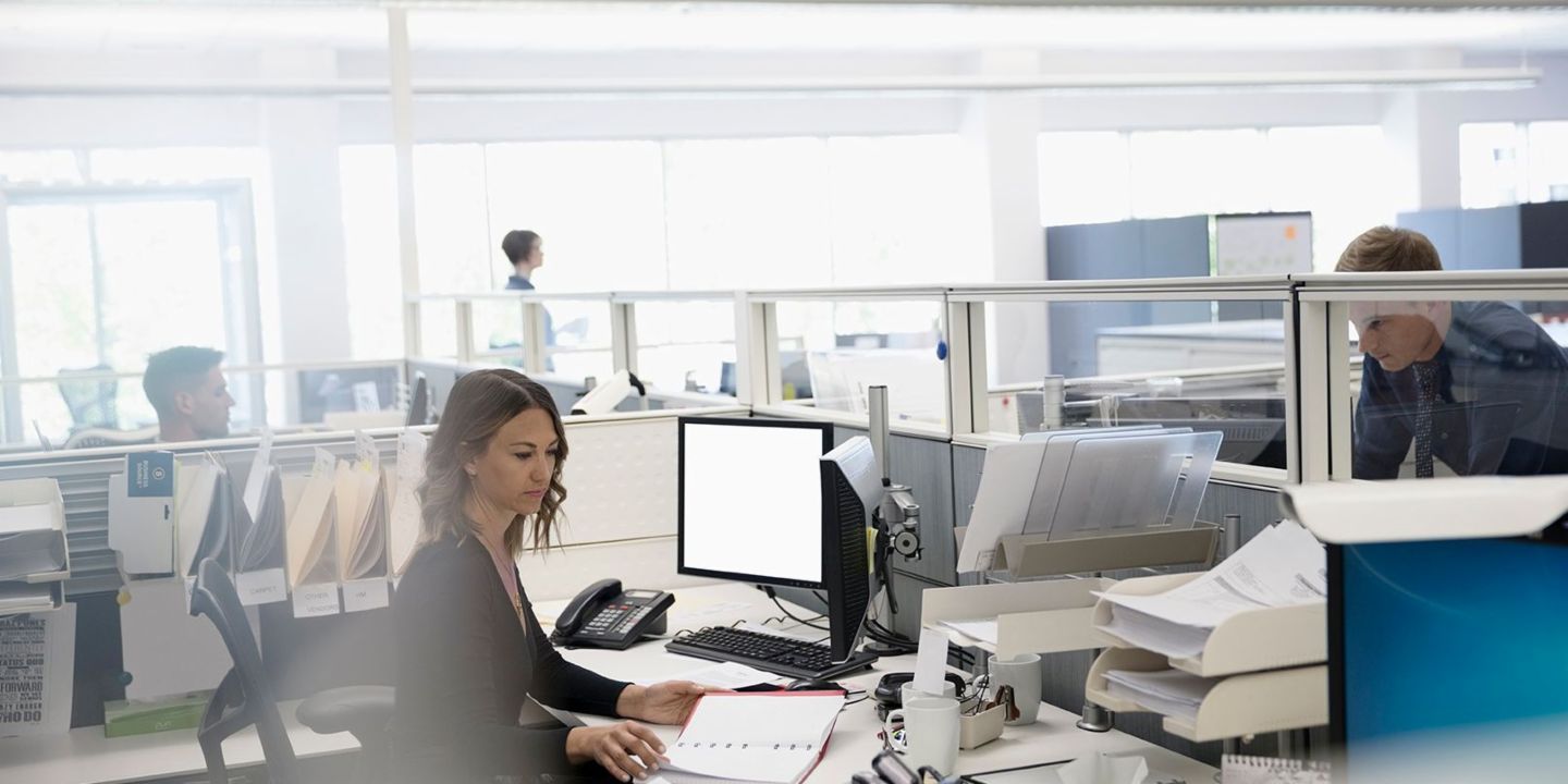 Дигитализацията: трима служители са в отворен офис – една жена е седнала на компютъра си измежду хартиите